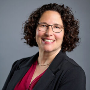 Smiling woman with short curly hair and glasses, wearing a dark blazer over a red blouse, posed against a plain gray background.