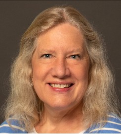 A woman with long, wavy, blonde hair and a striped shirt smiles at the camera against a dark background.
