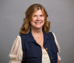 Woman with wavy blonde hair wearing a navy vest over a cream blouse, smiling at the camera against a plain gray background.