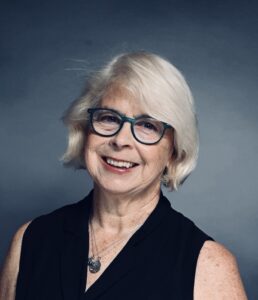 Woman with white hair and glasses, wearing a sleeveless black top and necklaces, smiles in front of a plain gray background.