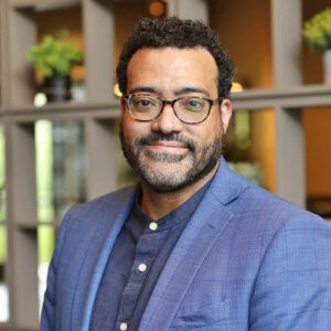 A man with a beard and glasses wearing a blue blazer over a button-down shirt.