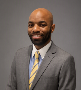 A man wearing a gray suit and yellow striped tie stands in front of a plain gray background, smiling at the camera.
