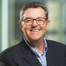 Smiling man wearing glasses, a dark blazer, and a checked shirt, standing indoors with a blurred background.