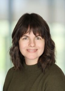 Woman with shoulder-length brown hair and bangs, wearing a green sweater, smiling in front of a blurred background.