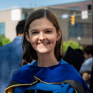 Woman in academic regalia smiles outdoors on a sunny day, with blurred people and buildings in the background.