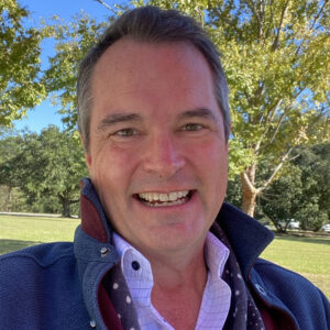 A man smiling outdoors in a park, wearing a blue jacket over a white shirt with a scarf, with green trees and blue sky in the background.