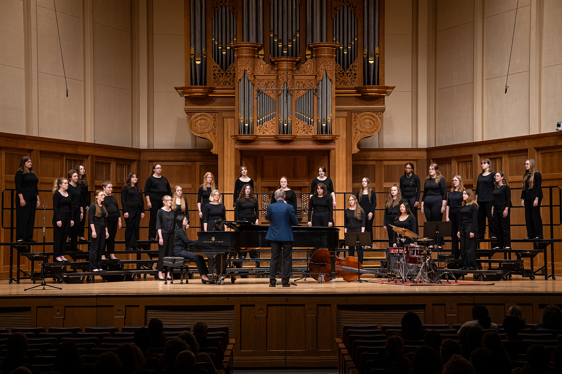 A conductor leads a choir of women dressed in black on stage, accompanied by a pianist, with an organ and drum set in the background.