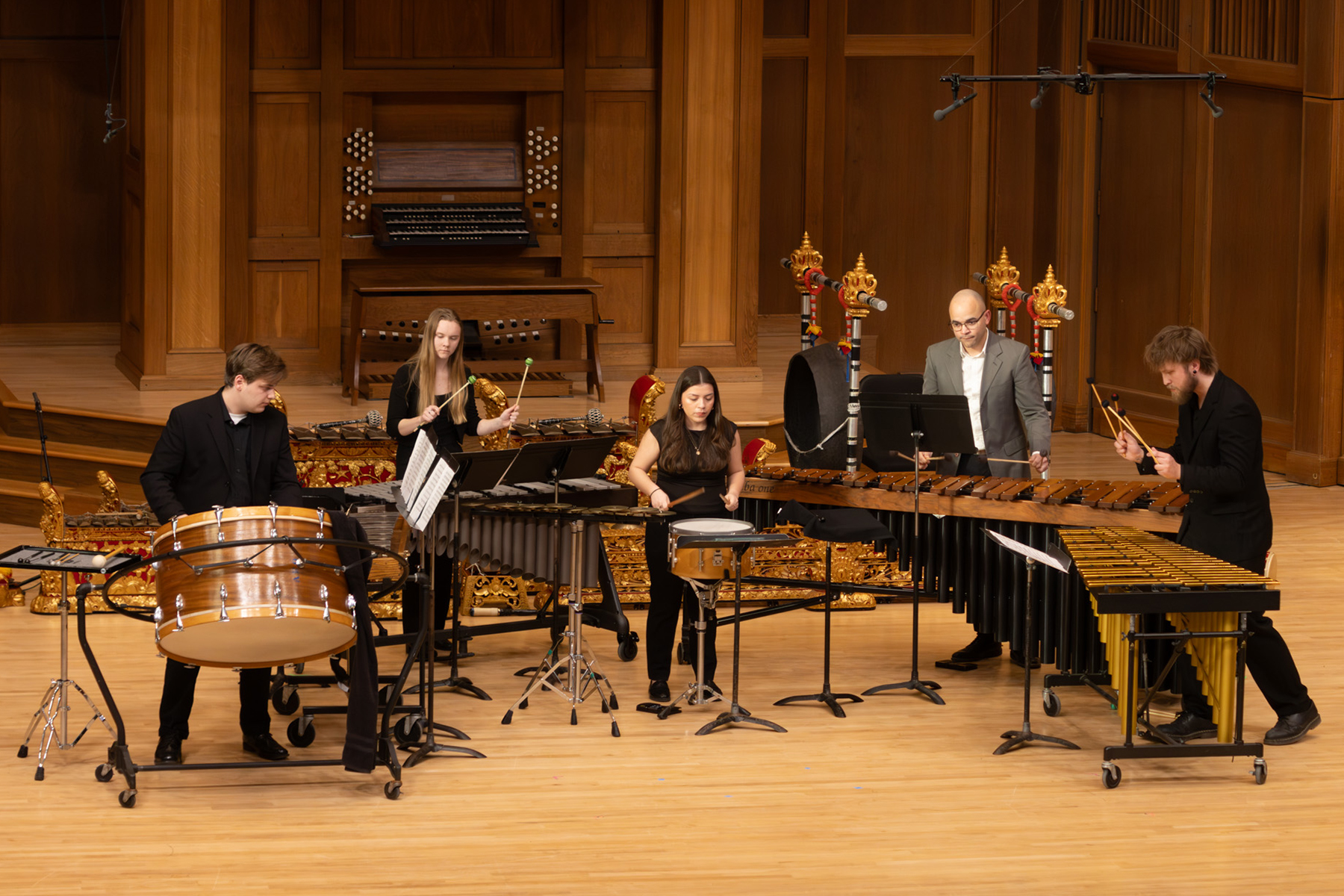 Five musicians perform on stage with percussion instruments, including drums, marimbas, and vibraphones, in a wooden concert hall.
