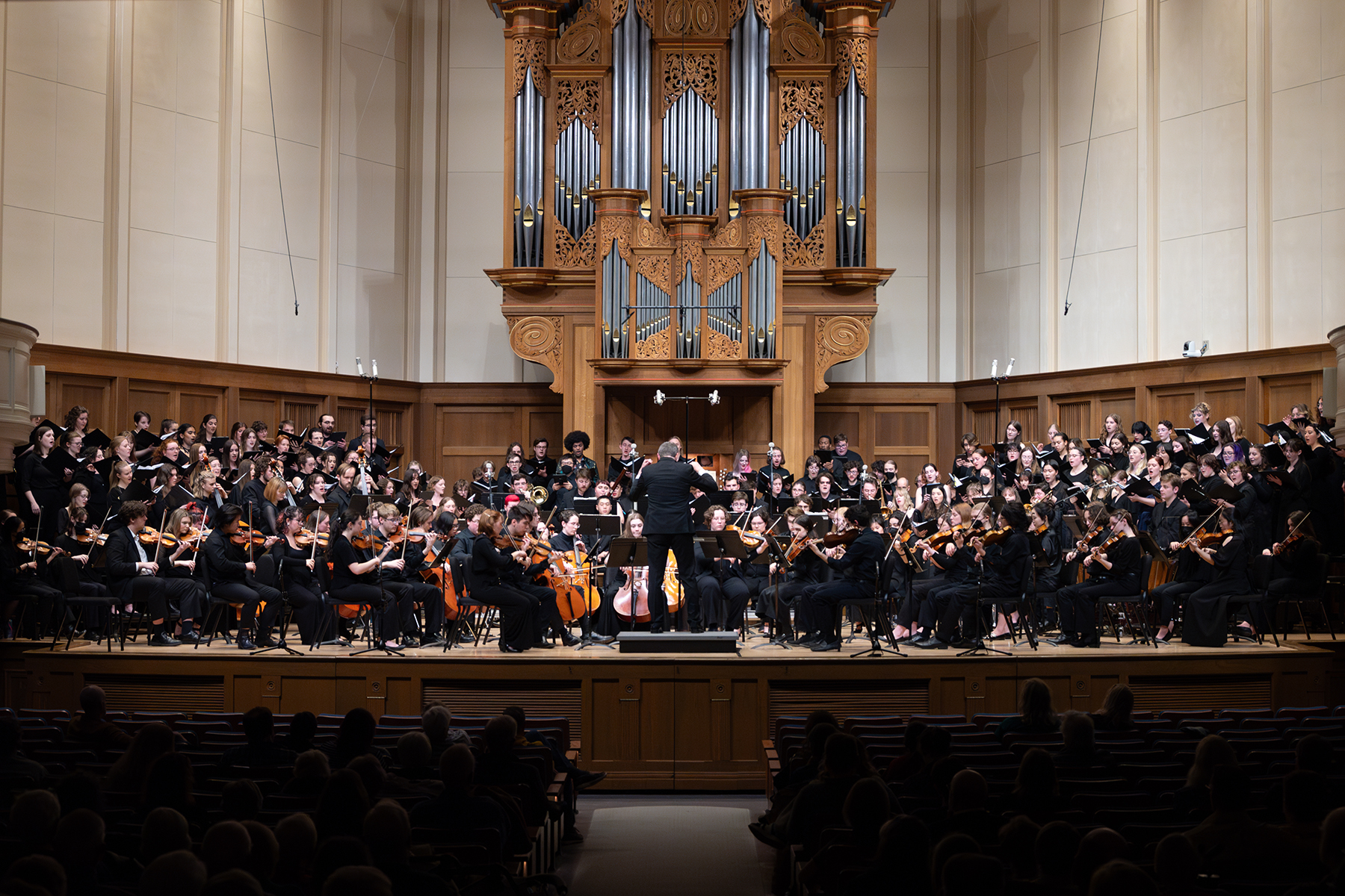 A conductor leads a large orchestra and choir onstage in a concert hall with a pipe organ in the background.