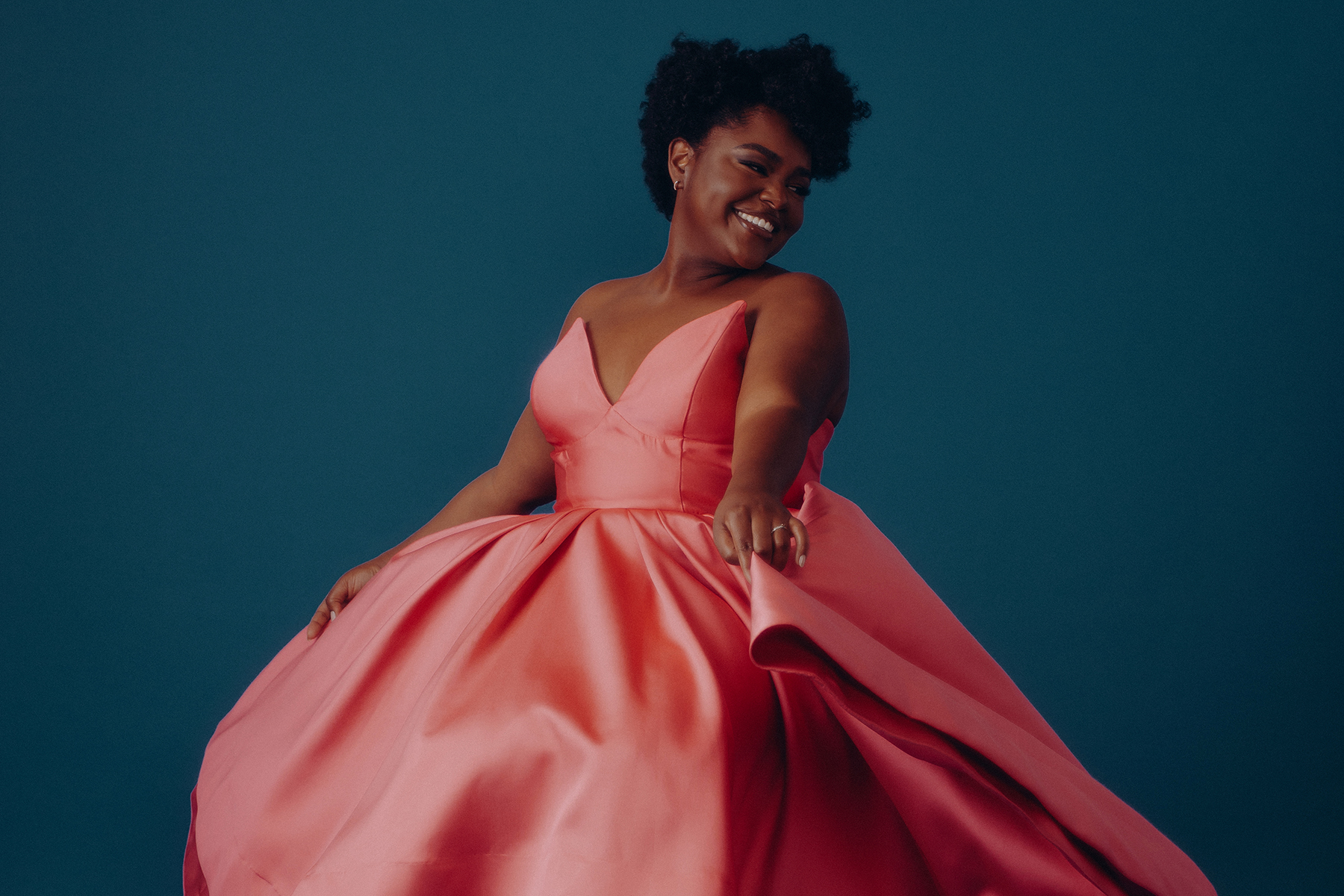 A woman in a pink, sleeveless gown stands against a blue background, smiling and looking to her left while holding her dress out to the side.