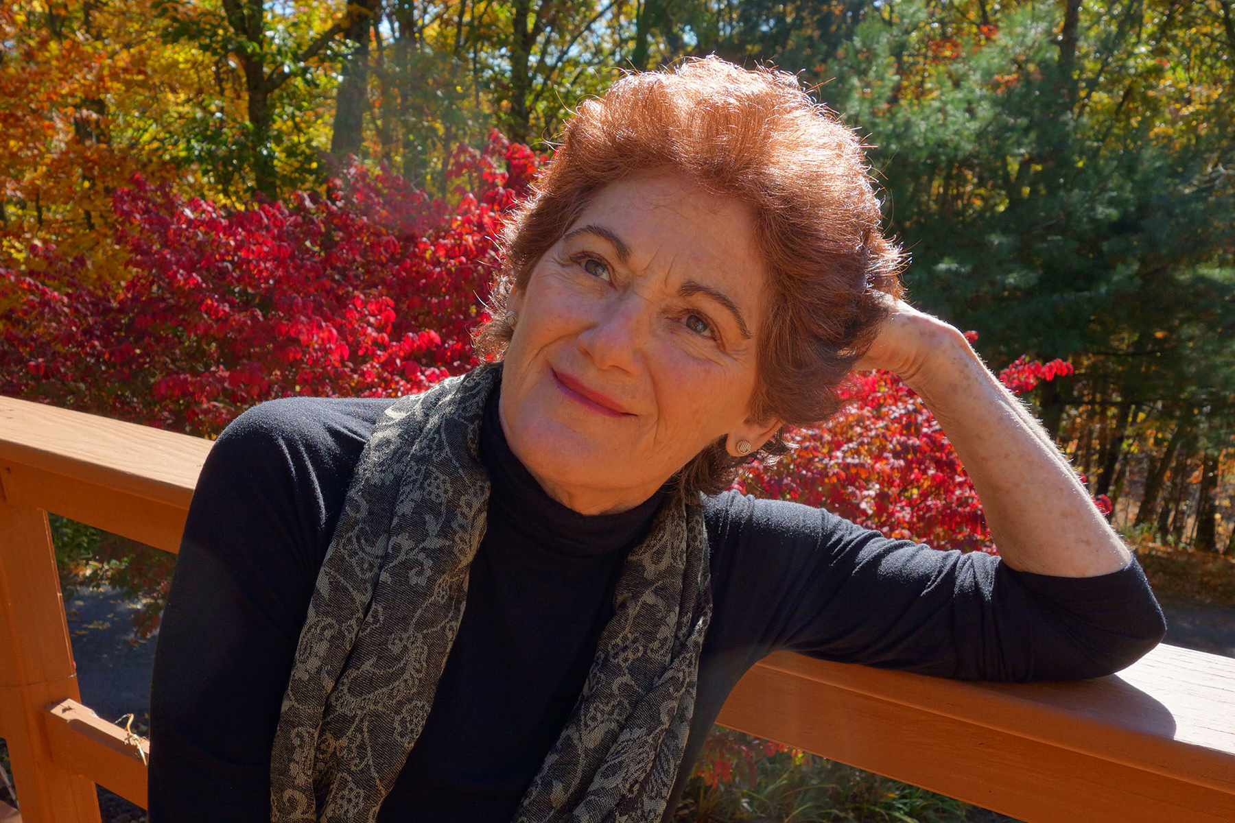 An older woman with short brown hair sits outdoors on a wooden deck, leaning on her arm, with red and green foliage in the background.