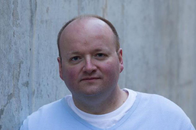 A man with a shaved head and light skin wears a pale blue shirt, standing in front of a concrete wall, looking at the camera.