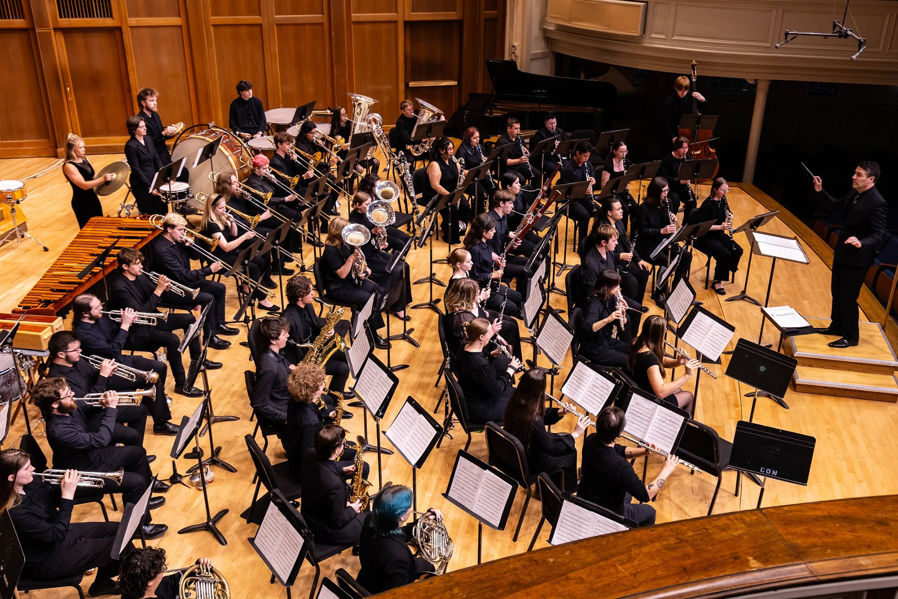 A large wind band dressed in black performs on stage with various instruments while a conductor leads them.