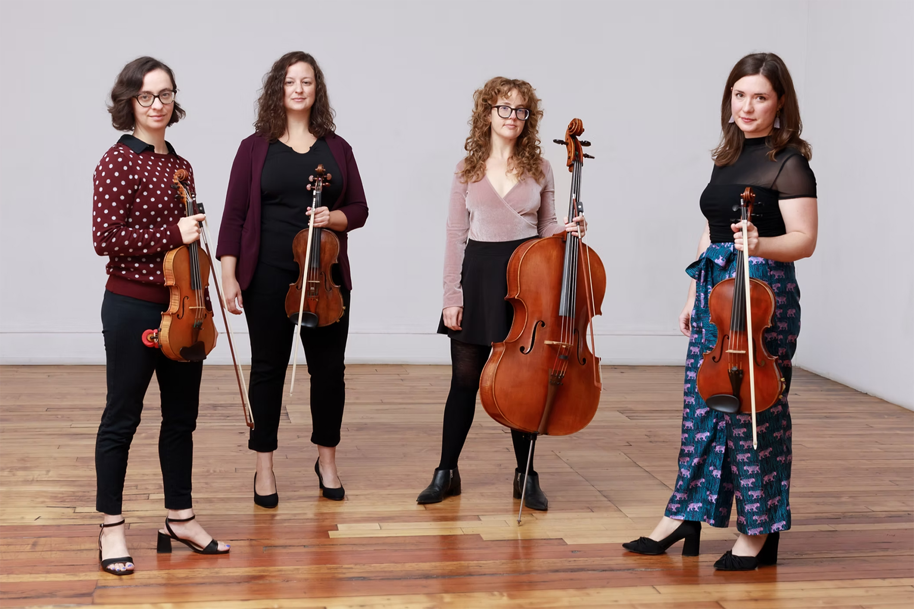 Four women stand indoors holding string instruments, including violins, a viola, and a cello, on a wooden floor with a plain gray wall behind them.