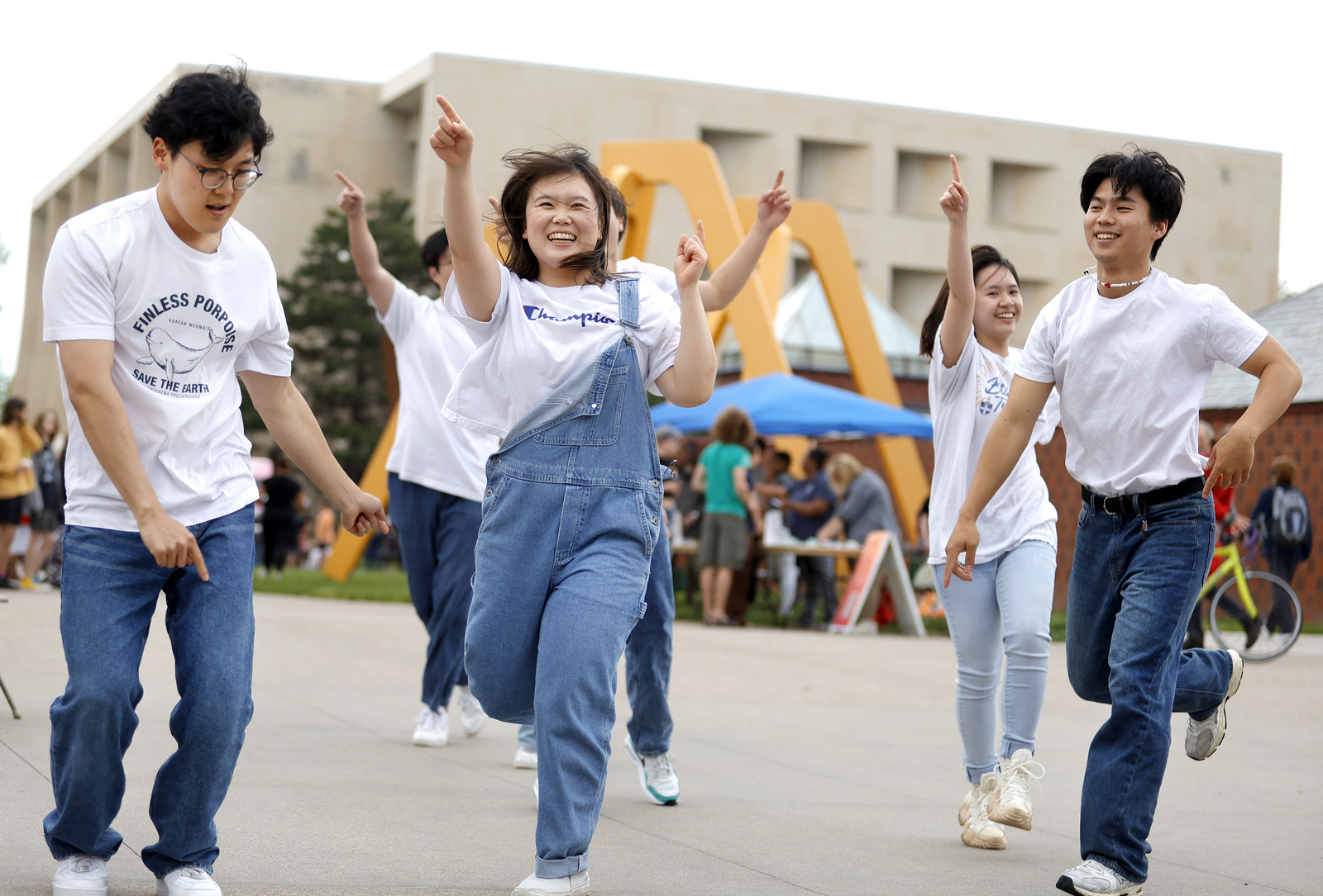 Five people dance energetically outdoors, smiling, with a crowd and modern building in the background.
