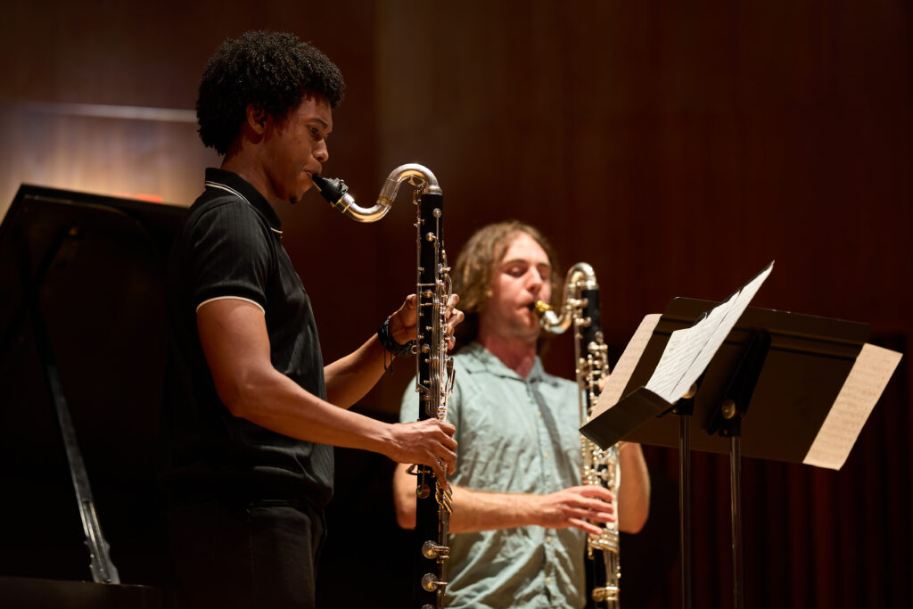 Two musicians play bass clarinets on stage, reading sheet music from a stand. A grand piano is visible in the background under warm lighting.