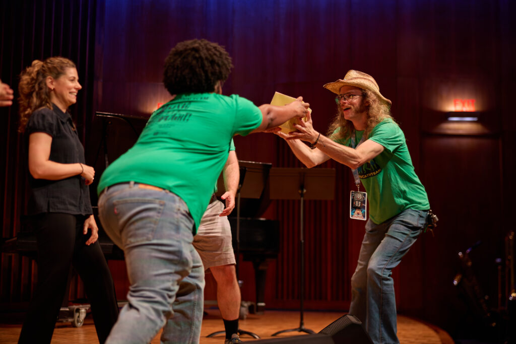 Two people in green shirts exchange a yellow folder on stage, while others stand nearby. The setting appears to be a music or performance venue with a piano in the background.