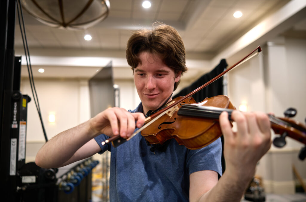 A young person in a blue shirt plays a violin indoors, focusing intently on the instrument under bright ceiling lights.