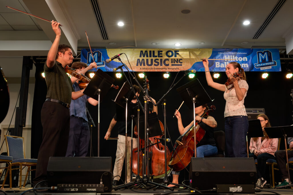 A small orchestra performs on stage under a Mile of Music Inc banner. Musicians play violins and a cello, with one standing woman raising her bow. Bright stage lights shine behind them.