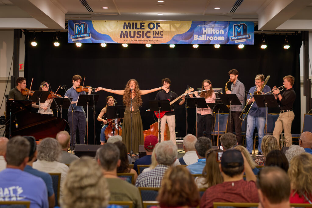 A band performs on stage with various instruments while a woman sings at the center, facing an audience seated in a ballroom during the Mile of Music event.