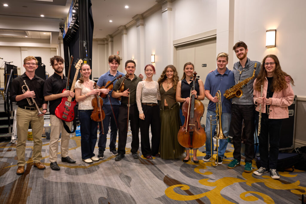 Eleven young adults stand indoors in a line, holding various musical instruments, including brass, strings, woodwind, and electric guitar, posing and smiling for a group photo.