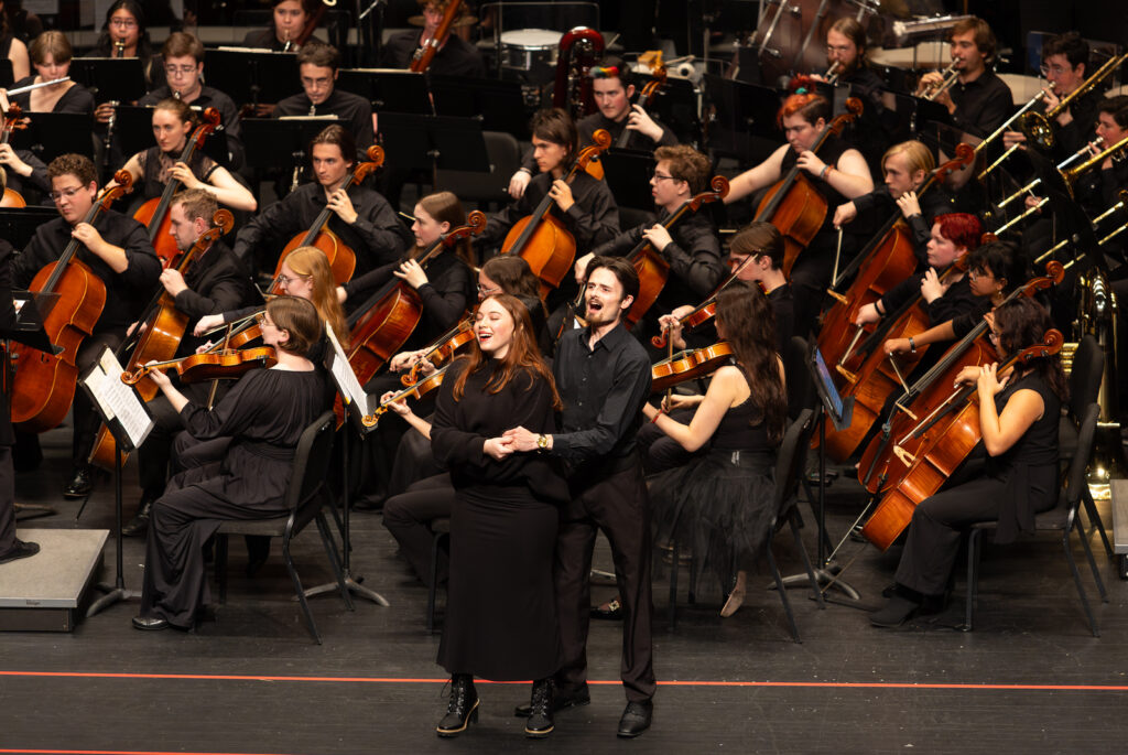 A large symphony orchestra performs on stage while a woman and a man stand at the front singing, holding hands.