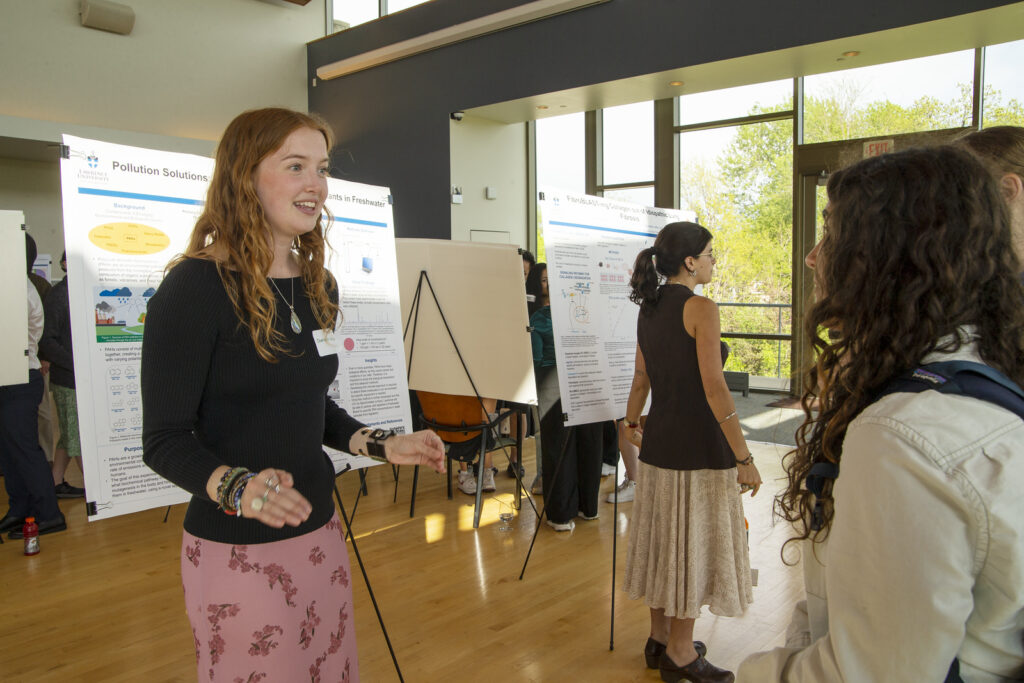 A woman presents a science poster to another attendee at an indoor event, with other people viewing posters in the background. Sunlight streams through large windows.