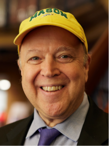 Smiling man wearing a yellow cap, dressed in a suit and tie, in an indoor setting.