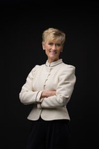 Person with short blond hair wearing a cream-colored jacket stands against a black background with arms crossed, smiling slightly.