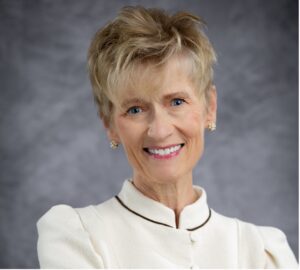 Smiling woman with short blonde hair, wearing a cream blazer, stands in front of a gray background.