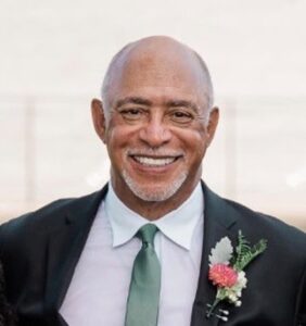 Smiling man in a suit and green tie, wearing a boutonniere with pink and white flowers, standing in front of a light, blurred background.