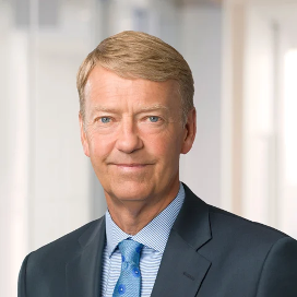 A man with short light brown hair in a suit and blue tie, standing in an office setting with blurred background.