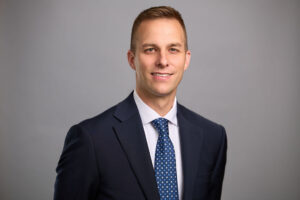 A man in a dark suit and blue patterned tie poses against a plain gray background, smiling at the camera.