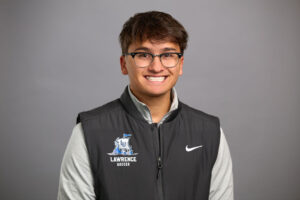 A person wearing glasses, a gray long-sleeve shirt, and a black Lawrence Soccer Nike vest poses in front of a plain gray background, smiling.