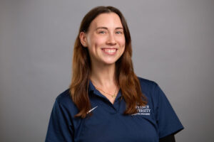 Woman with long brown hair wearing a navy blue Lawrence University polo shirt stands against a plain gray background, smiling at the camera.
