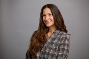 Woman with long brown hair, wearing a plaid blazer and blue top, smiles at the camera against a plain gray background.