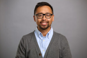 A man with short dark hair, glasses, and a beard is wearing a light blue shirt and gray cardigan. He is smiling and standing in front of a plain gray background.