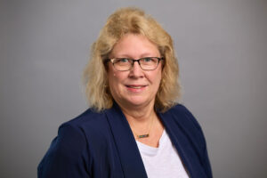 Woman with blonde hair and glasses wearing a dark blue blazer and white top, smiling at the camera against a plain light gray background.