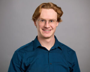 A person with light skin, short wavy hair, and glasses is smiling at the camera, wearing a navy button-down shirt against a plain gray background.