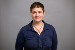 Person with short hair and a navy blue shirt stands against a plain gray background, facing forward and smiling.