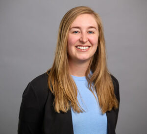 A person with long blonde hair, wearing a light blue top and black blazer, smiles while facing the camera against a plain gray background.