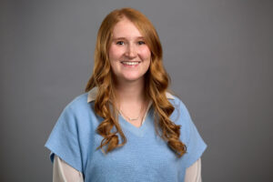 Person with long red hair wearing a blue sweater over a white shirt stands smiling in front of a plain gray background.