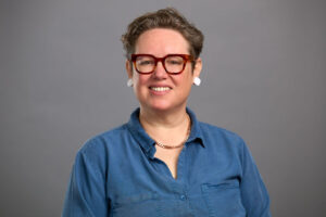 Person with short hair, glasses, and earrings wearing a blue button-up shirt and necklace, smiling in front of a plain gray background.