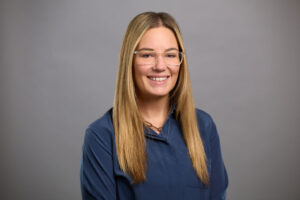 Woman with long straight blonde hair and glasses, wearing a blue blouse, smiling in front of a plain gray background.