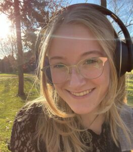 A person with long blonde hair, glasses, and headphones smiles outdoors in sunlight, with trees and grass in the background.