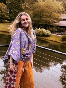 A person stands on a bridge over a pond, smiling, wearing a purple patterned top and orange pants with trees and a building in the background.