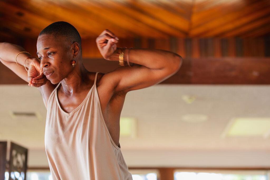 Person in a sleeveless shirt raises their arms while dancing indoors, with natural light and wooden ceiling visible in the background.