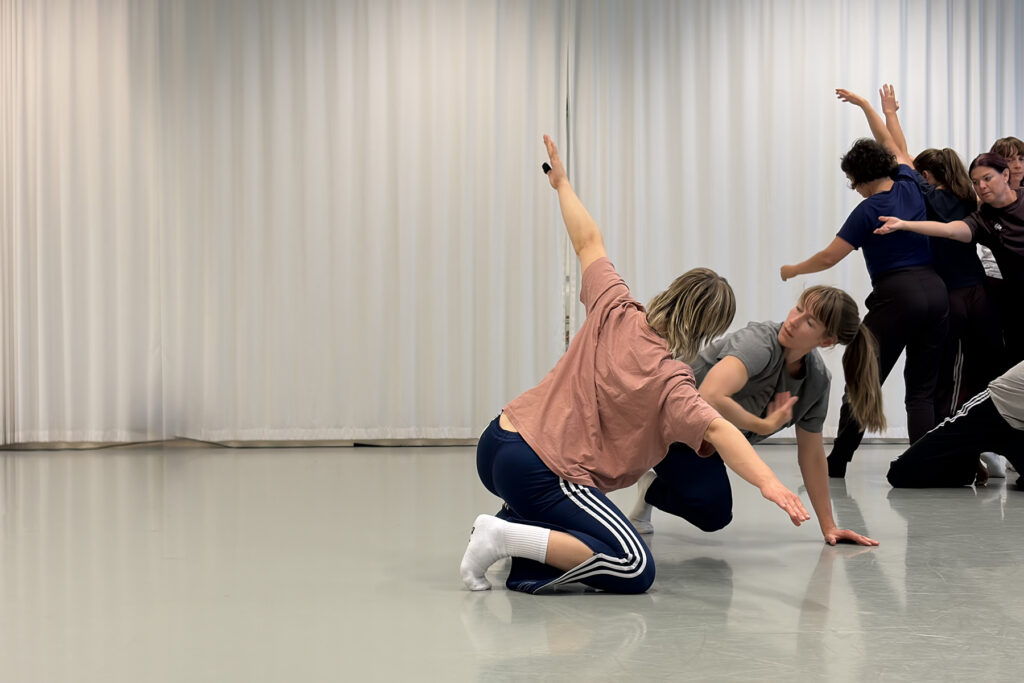Several people perform contemporary dance moves in a studio with a light floor and curtain backdrop, two kneeling in the foreground and others standing behind them.