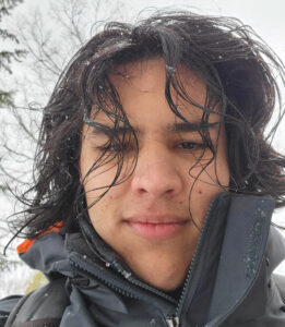 A person with long dark hair stands outdoors in winter, wearing a jacket.