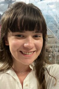 A person with long brown hair and bangs, wearing a white collared shirt, smiles at the camera.
